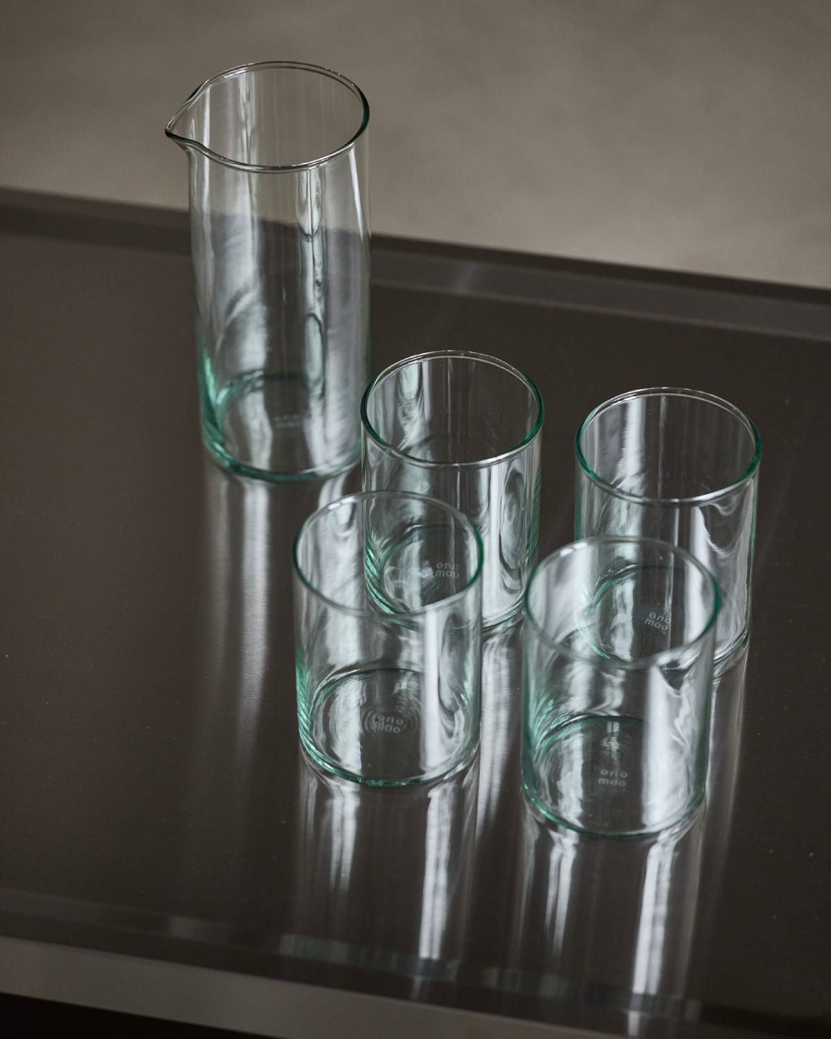 Vier hohe Trinkgläser und eine Karaffe aus grünlichem Glas mit onomao-Prägung auf einer dunklen Fläche.