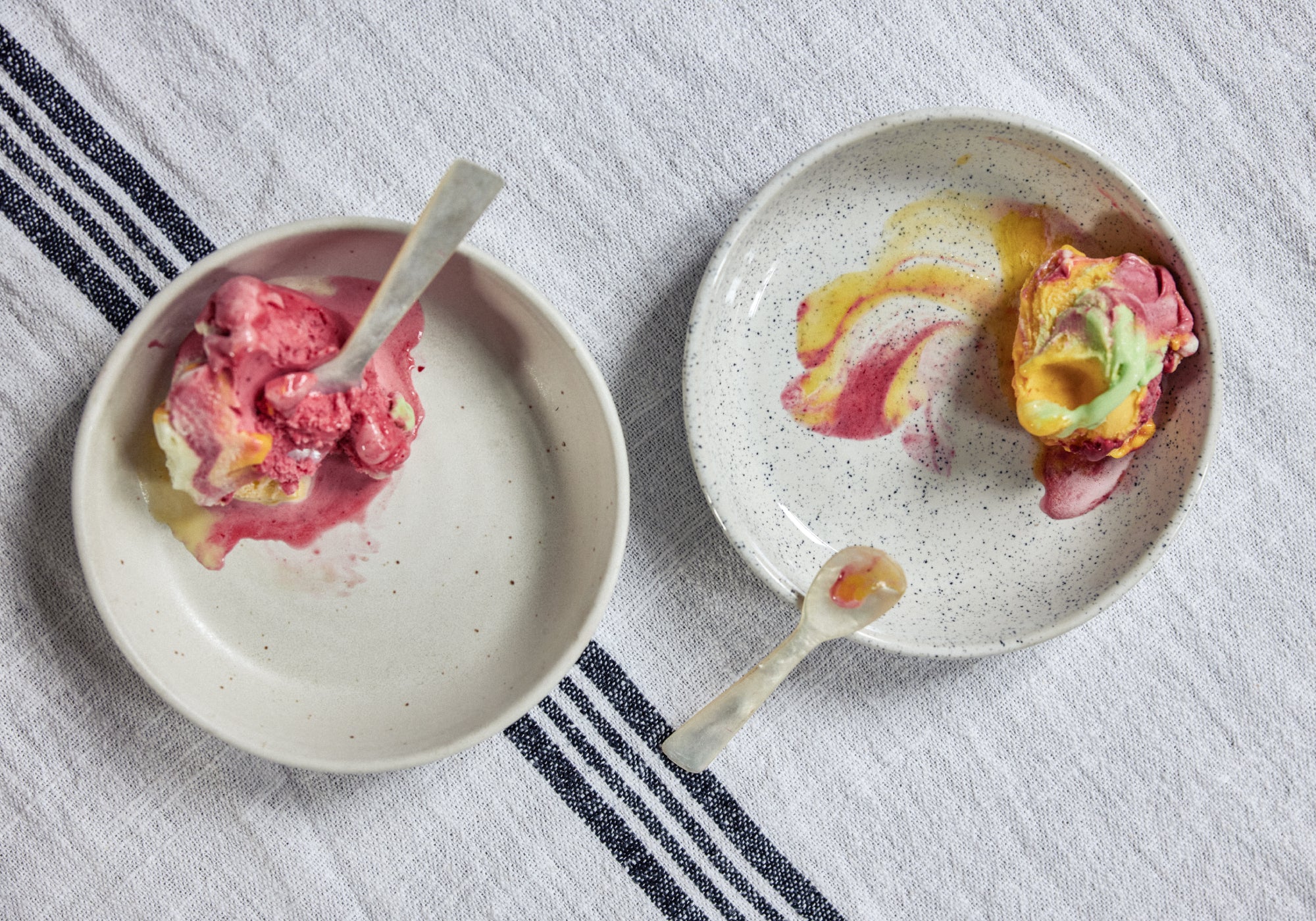 Zwei gesprengte Keramikschalen mit jeweils einer Kugel buntem Speiseeis in Pink, Gelb und Grün, das leicht angeschmolzen ist und bunte Schlieren hinterlässt. Dazu je ein heller Keramiklöffel, alles auf einem grau-weiß gestreiften Leinentuch von oben fotografiert.