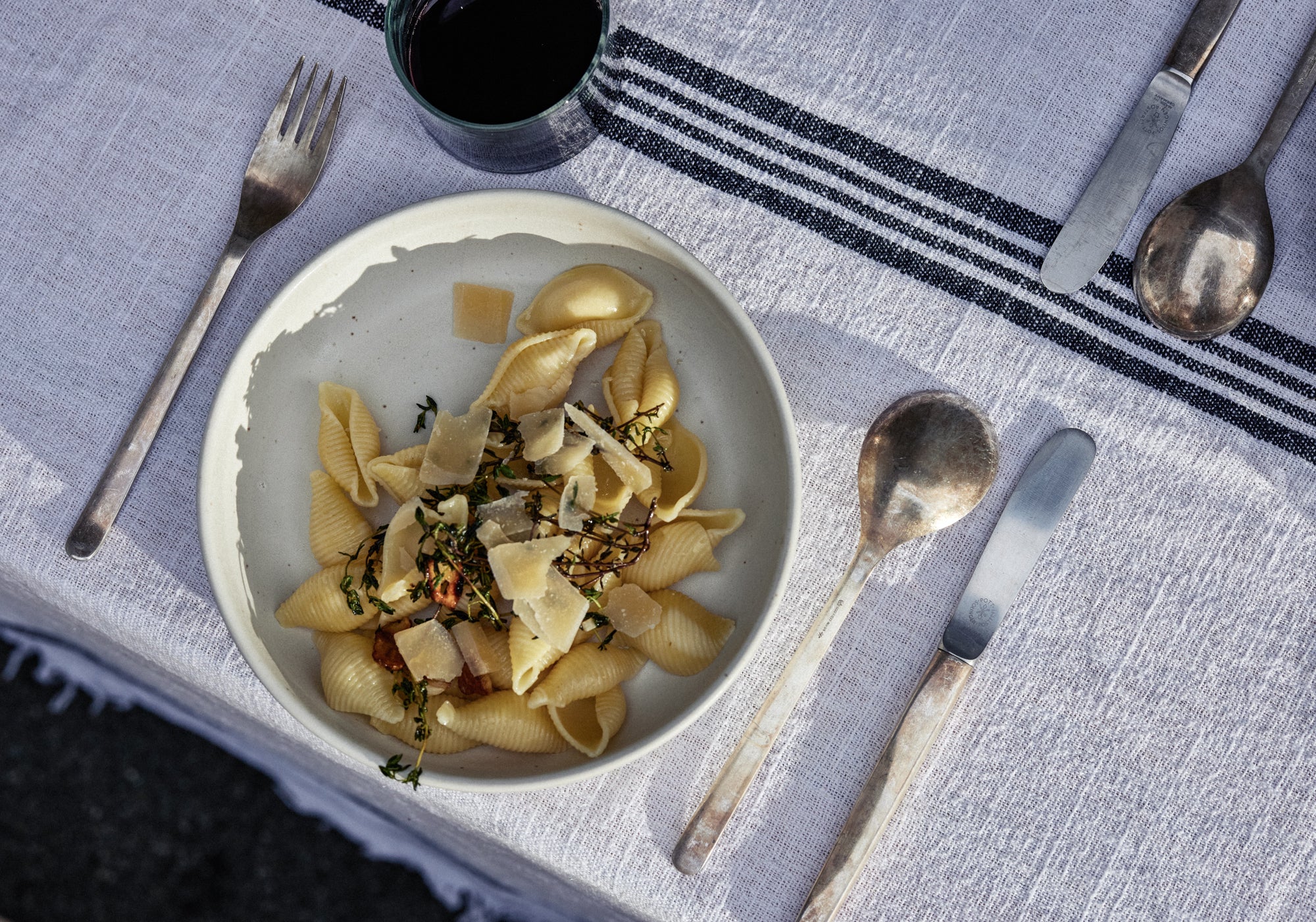 Cremefarbene Keramikschale mit Conchiglie-Pasta, Parmesanspänen und frischem Thymian auf einem hellgrauen Leinentischtuch mit dunklen Streifen. Daneben silbernes Besteck (Gabel, Messer und Löffel) sowie ein dunkler Keramikbecher im Hintergrund.