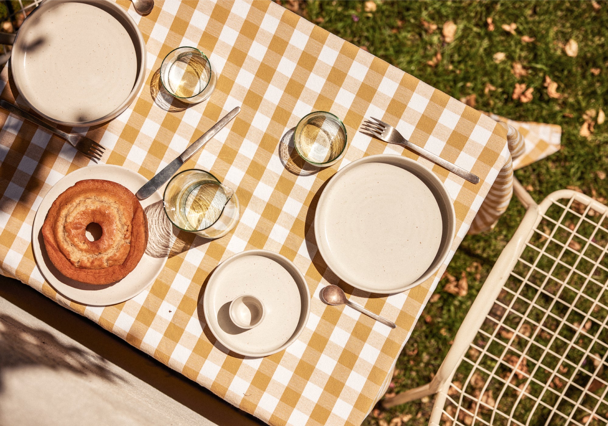Gartentisch von oben mit ockergelb-weiß kariertem Tischtuch, darauf cremefarbenes Keramikgeschirr (Teller, Schalen und ein kleiner Zuckertopf), silbernes Besteck und drei Wassergläser. Links ein goldbrauner Gugelhupf auf einem Teller. Rechts ein weißer Metallgartenstuhl, im Hintergrund grüner Rasen mit Herbstlaub.