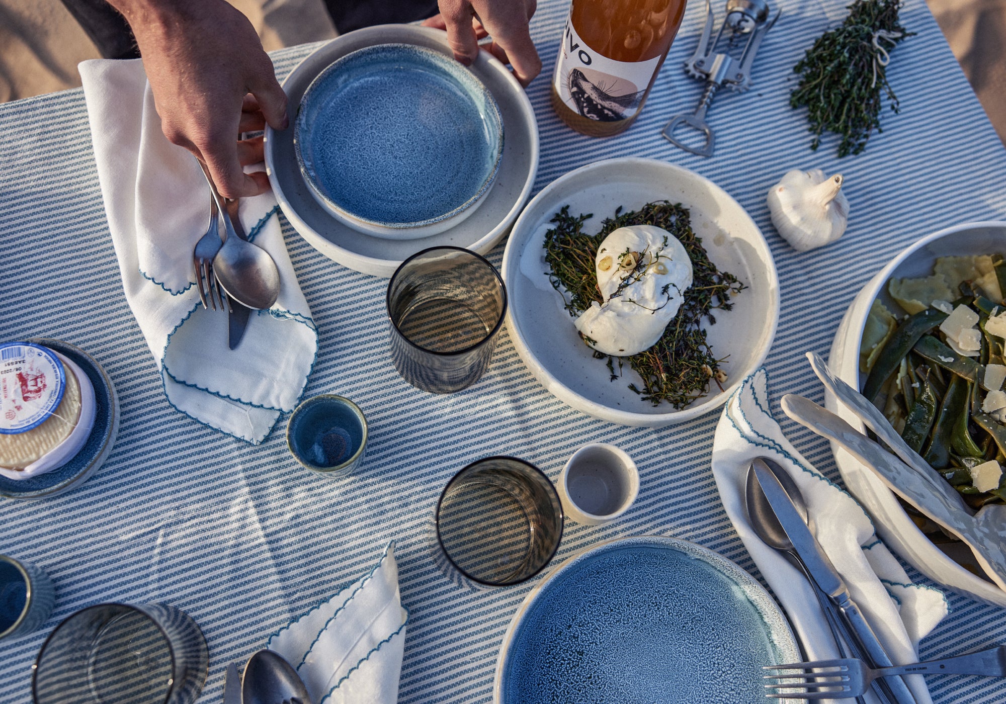Gedeckter Tisch von oben mit hellblau gestreiftem Leinentischtuch: Hände reichen blaue Keramikteller, daneben eine weiße Servierschale mit Burrata auf geröstetem Thymian, gemusterте Gläser, weiße Stoffservietten mit blauer Stickerei, silbernes Besteck, eine Knoblauchknolle, frische Kräuter, ein Käse sowie eine Weinflasche mit Korkenzieher.