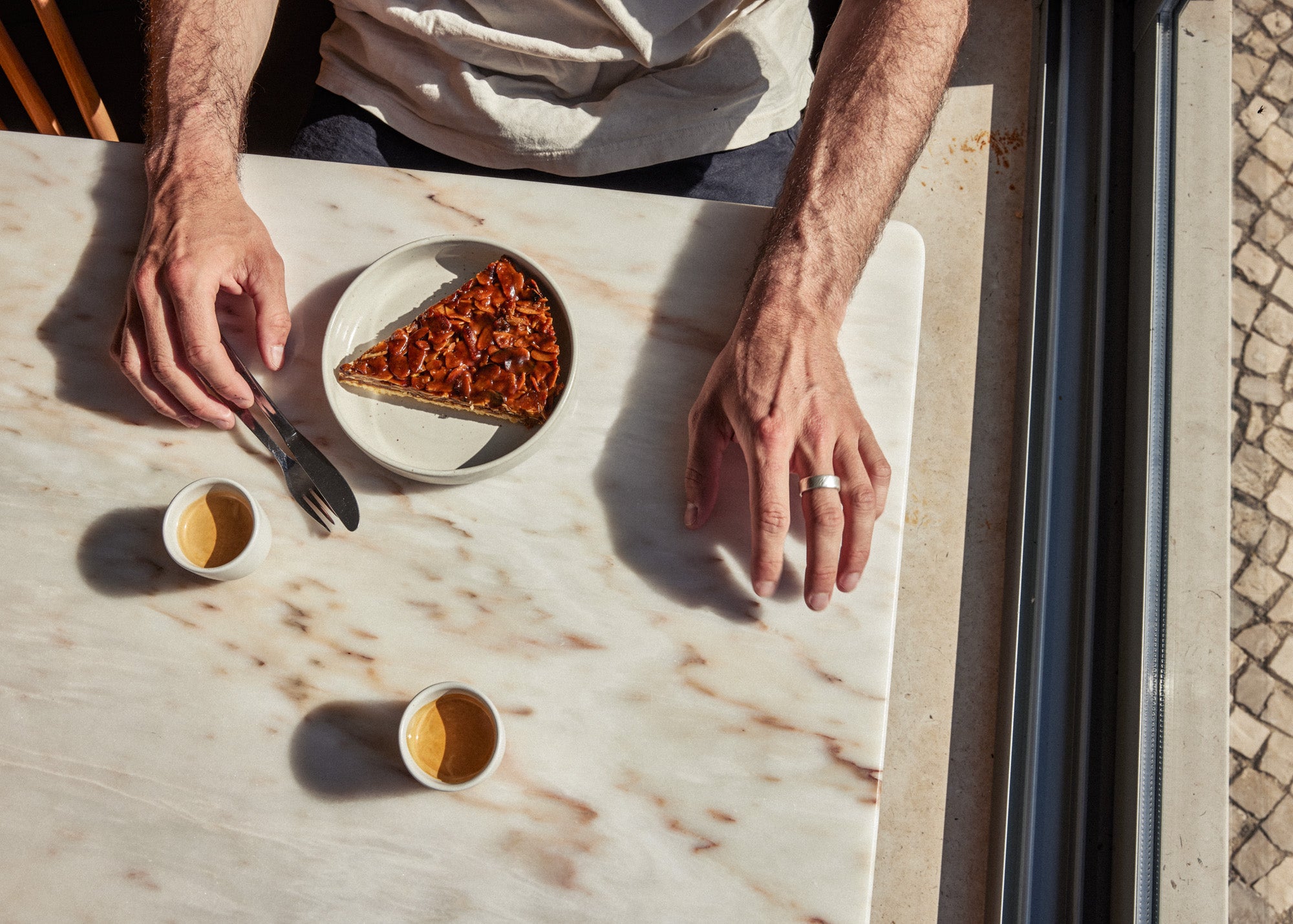 Hände einer Person greifen nach Besteck neben einem Stück Nuss-Karamell-Tarte auf einem cremefarbenen Keramikteller sowie zwei kleinen Espressotassen – alles auf einem Marmortisch von oben fotografiert im Sonnenlicht.