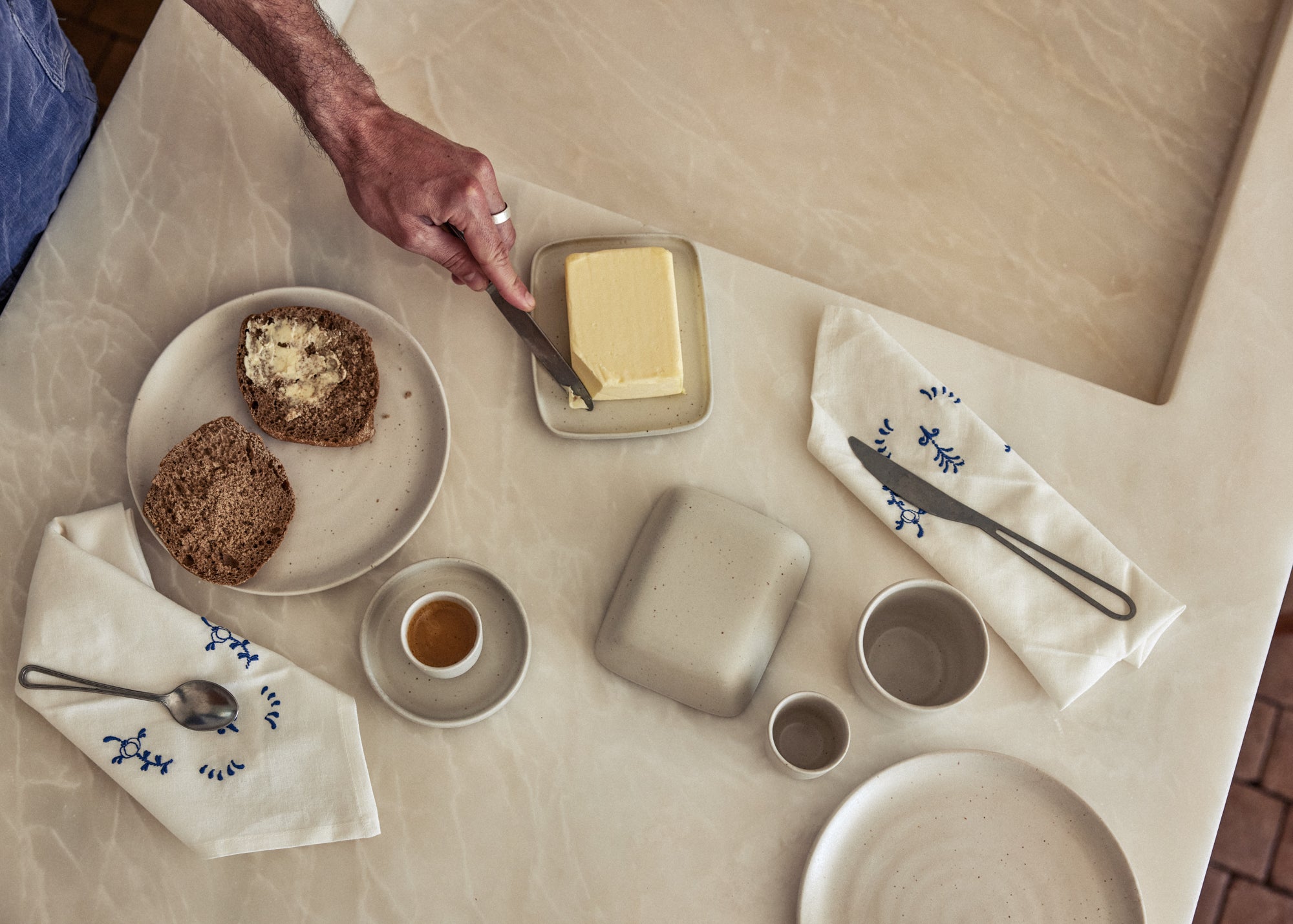 Vogelperspektive auf einen hellen Frühstückstisch. Eine Hand mit Ring streicht Butter auf Körnerbrot, das auf einem gesprenkelten Keramikteller liegt. Daneben eine Keramik-Butterdose mit frischer Butter, eine kleine Espressotasse auf Untertasse, eine geschlossene quadratische Keramikdose sowie kleine Becher und ein Teller in gleichem Naturton. Zwei weiße Leinenservietten mit blauer Blumenstickerei und je einem Besteckteil rahmen die Szene ein